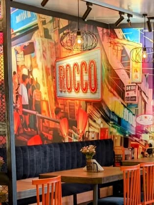 A painted Bangkok street scene with the Rocco sign glowing over the shopfronts
